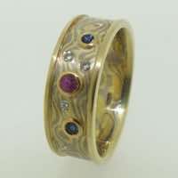 18k white and yellow gold mokume gane flat band with raised polished rails and flush set diamond. Set with blue sapphires and a ruby in low 22k gold rub-over settings.