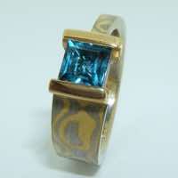 18k white and yellow gold mokume gane tapered band with a square facet cut blue topaz in an 18k yellow gold double bar setting.