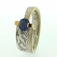 18k white gold and silver mokume gane tapered band with a blue sapphire in a 22k gold bar setting.  With flush set diamonds and a 22k gold flush rail detail.