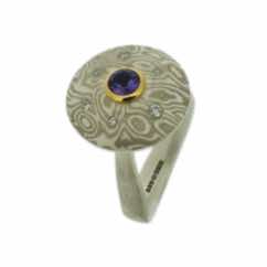 18k white gold and silver mokume gane discus with lilac sapphire in a 22k gold setting and diamonds presented on a sterling silver curved band.